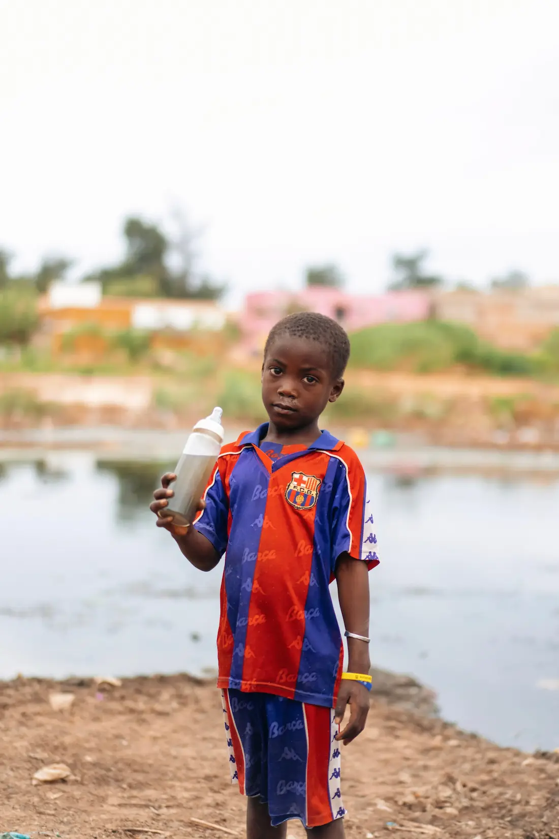 Un enfant tenant un biberon rempli d'eau trouble, debout près d'un plan d'eau